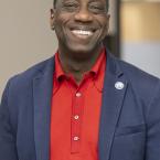 Dr. Julius Sokenu smiling, wearing a red shirt and blue sport coat with an MC Pin